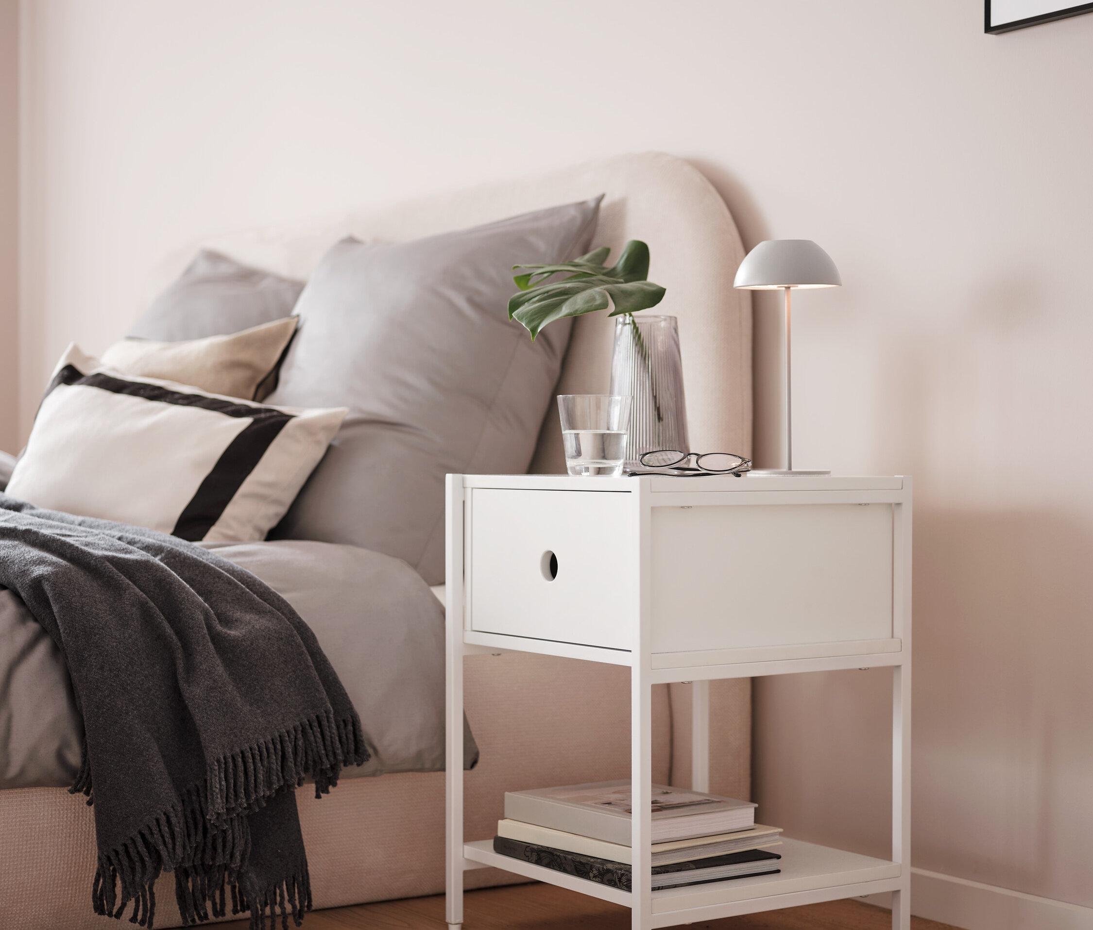 Šedá bateriová stolní lampa s LED na bílém nočním stolku vedle postele.