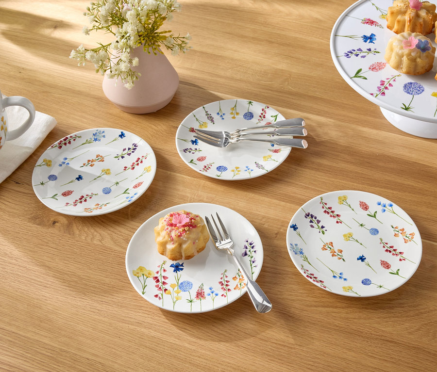 Na dřevěném stole je šálek s kávou, váza s květinami, talíře a dortíky.