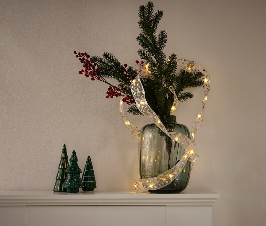 Váza s větvemi jehličnanů, červenými bobulemi a světelným řetězem se zlatými hvězdami. Vlevo stojí tři zelené keramické stromečky.