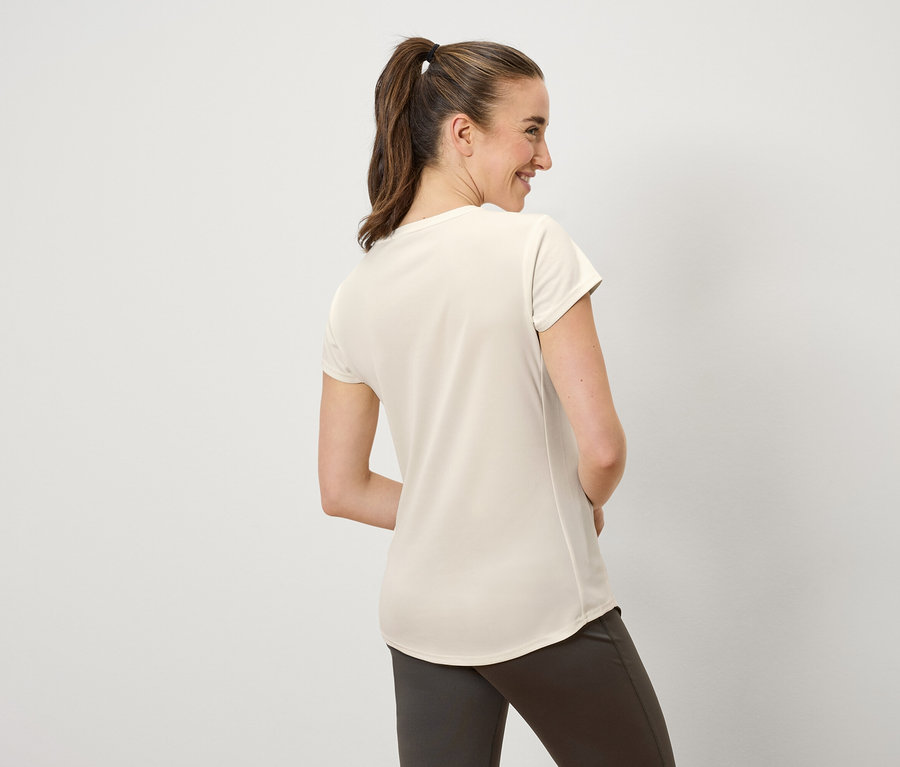 Žena s hnědými vlasy staženými do culíku se usmívá a dívá se doleva. Má na sobě krémové tričko a hnědé kalhoty.