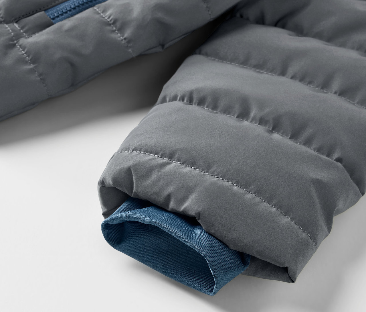 Detail šedé prošívané bundy s modrým zipem a lemem rukávu. Dětská reflexní prošívaná bunda.