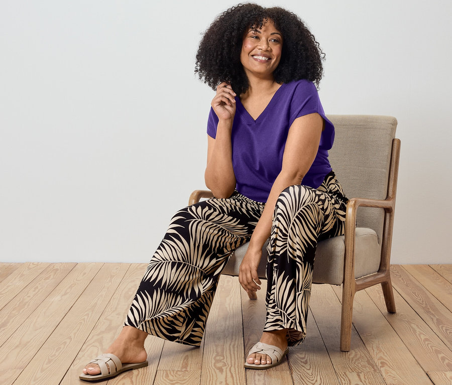 Žena sedí na židli a má na sobě fialový svetr a vzorované palazzo kalhoty.