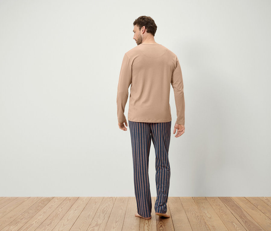Muž v béžovém tričku s dlouhým rukávem a modro-šedých pruhovaných kalhotách stojí zády ke kameře.