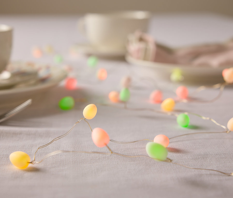 Detailní záběr stolu s nádobím a světelným řetězem ve tvaru velikonočních vajíček v pastelových barvách.