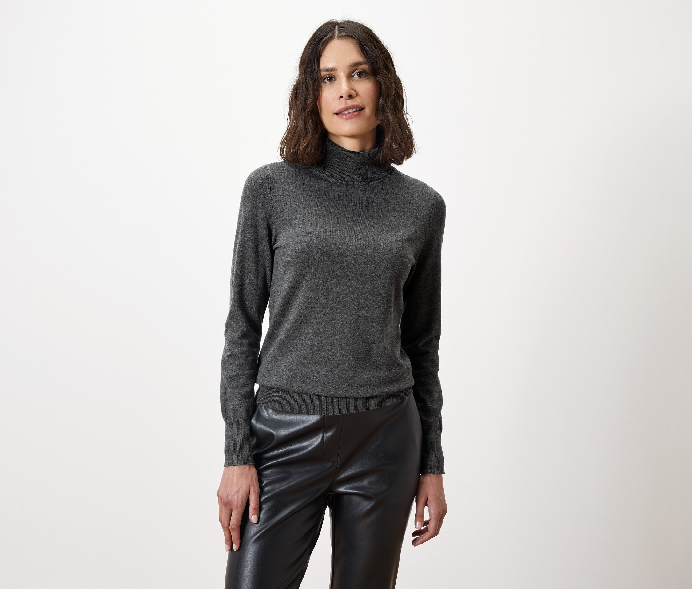 Žena pózuje v šedém roláku a černých kožených kalhotách.