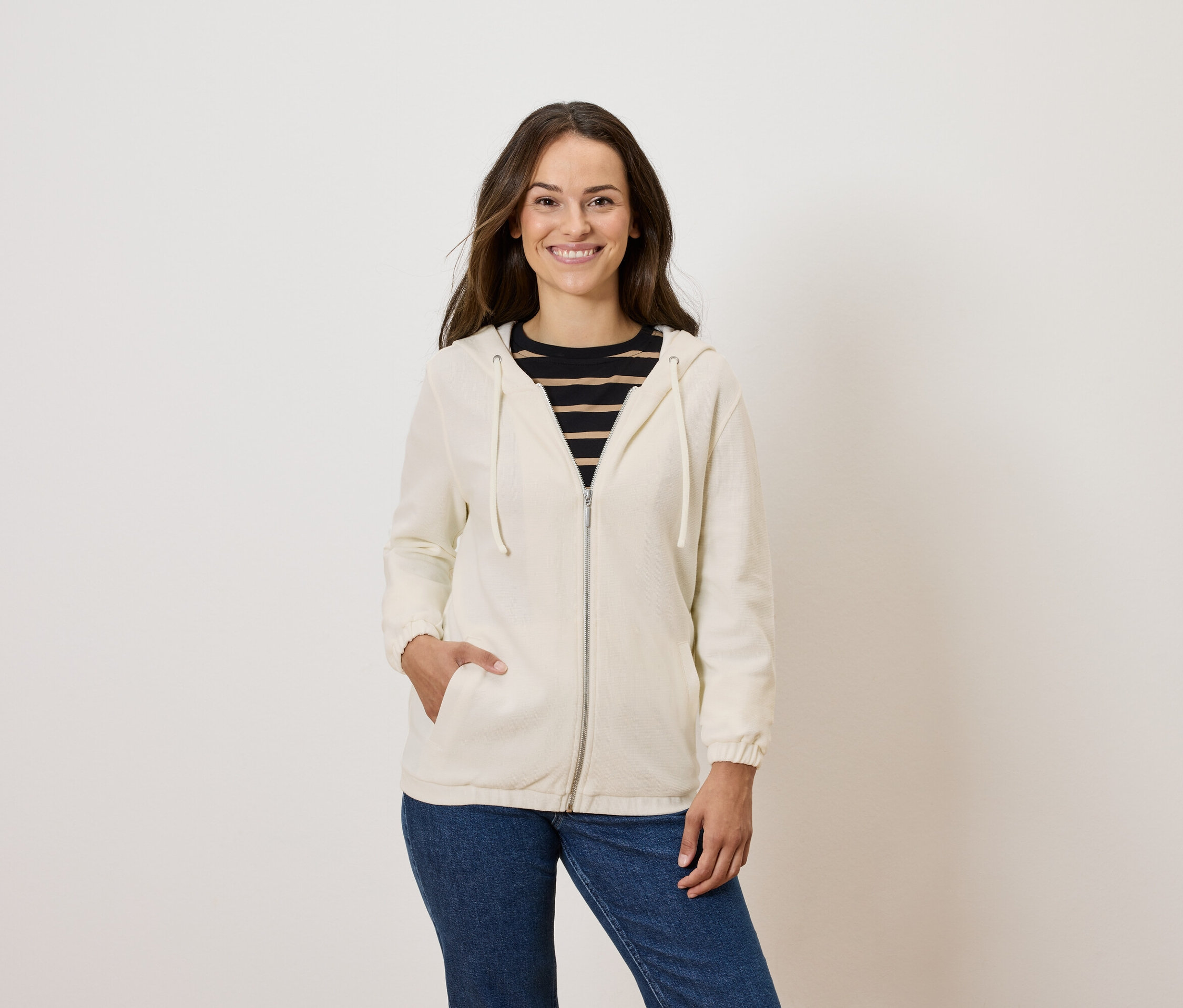Žena pózuje v krémové mikině na zip a modrých džínách.