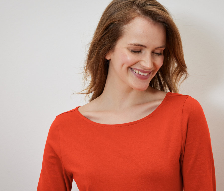 Usmívající se žena s dlouhými vlasy v oranžovém tričku s dlouhým rukávem.