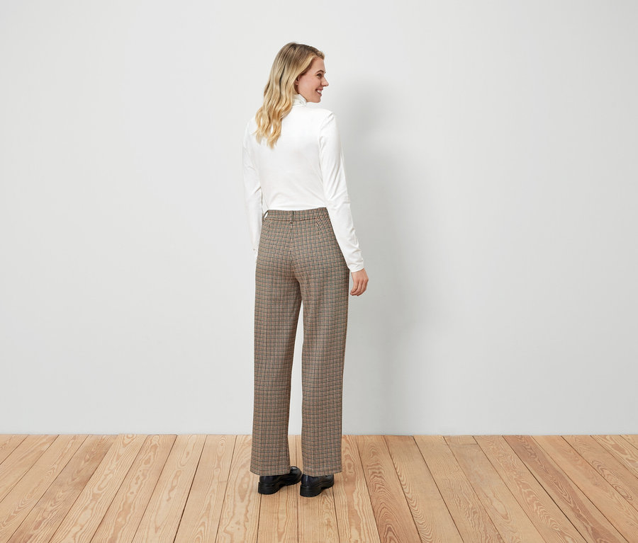 Žena pózuje v bílém topu a hnědých kostkovaných kalhotách.