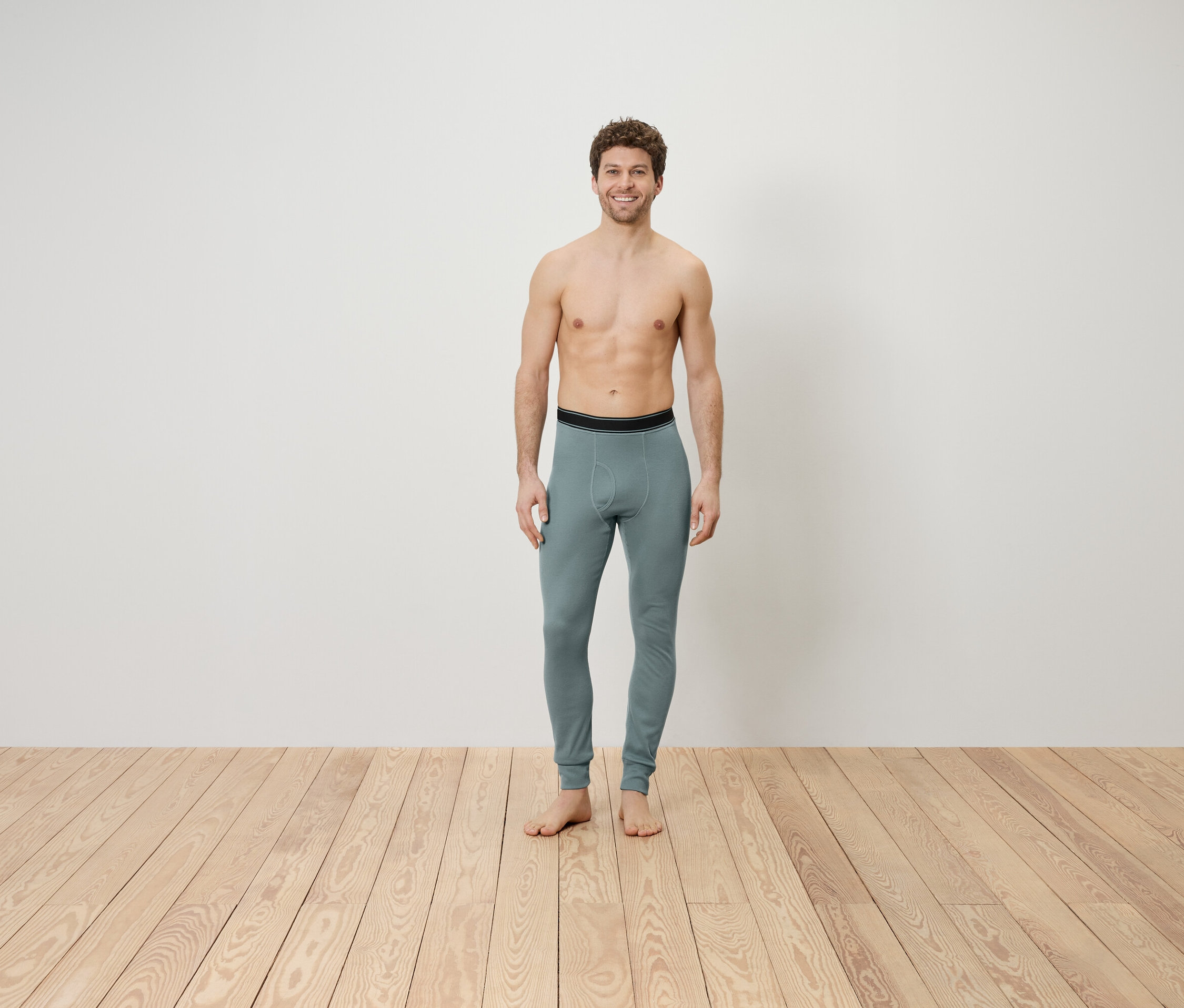 Muž s odhalenou horní částí těla stojí na dřevěné podlaze a má na sobě šedé legíny.