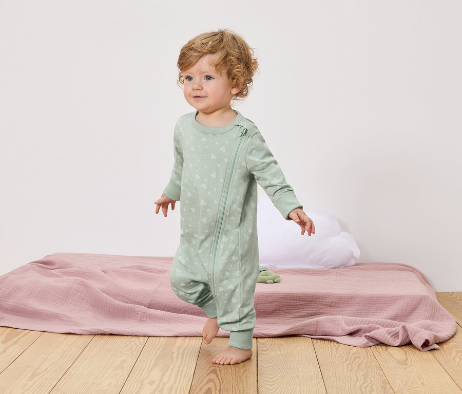 Malé dítě s kudrnatými blond vlasy chodí bosé po dřevěné podlaze. Má na sobě světle zelené pyžamo se zipem a vzorem srdce.
