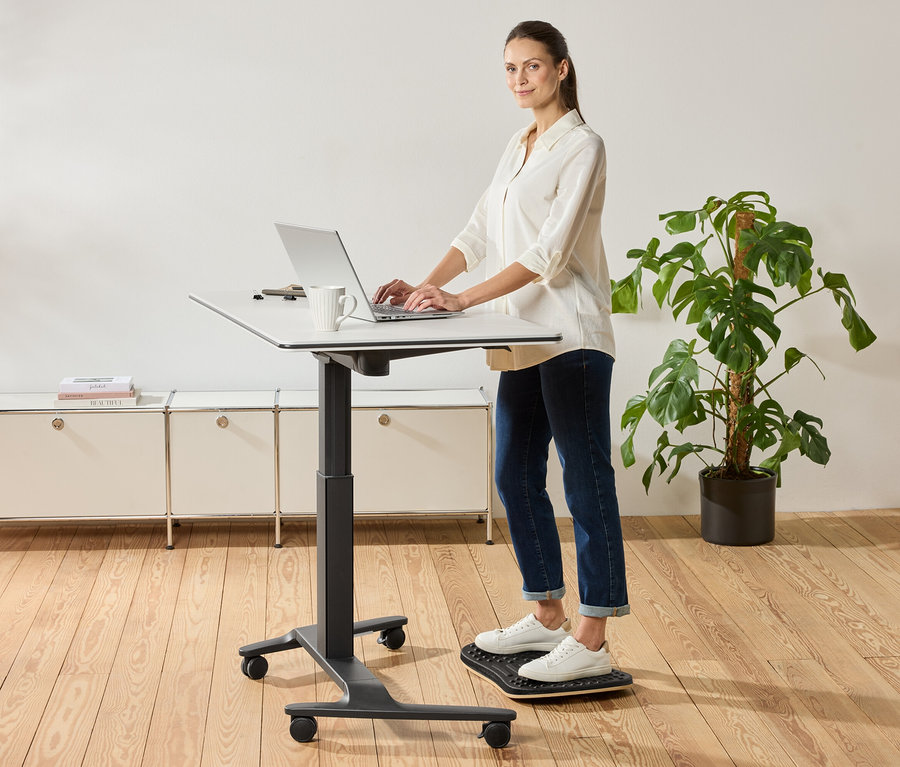 Žena stojí u výškově nastavitelného stolu a používá notebook, vedle stojí hrnek.