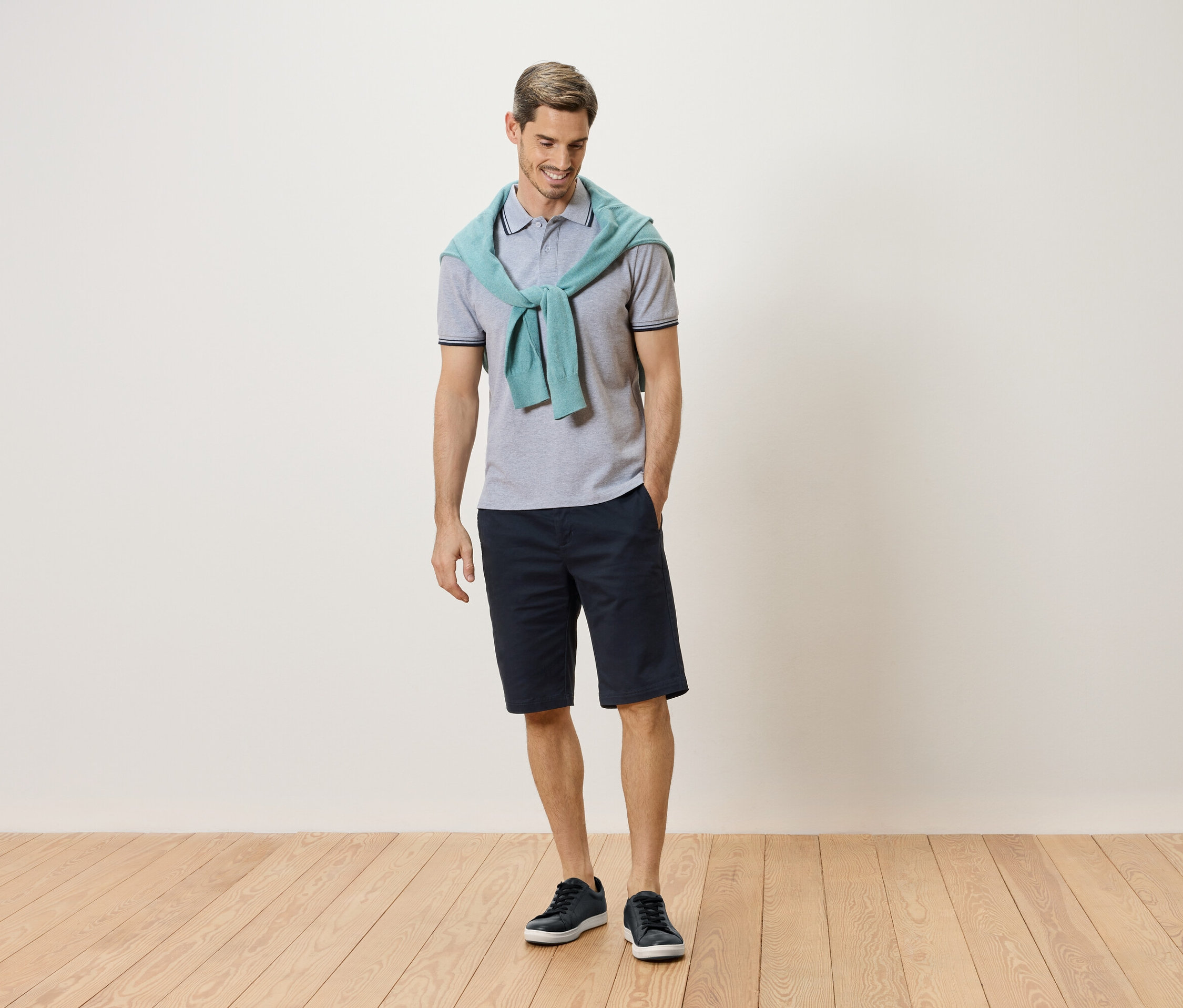 Muž stojí se světle modrým svetrem kolem krku, šedým polo tričkem, tmavě modrými šortkami a modrými teniskami.