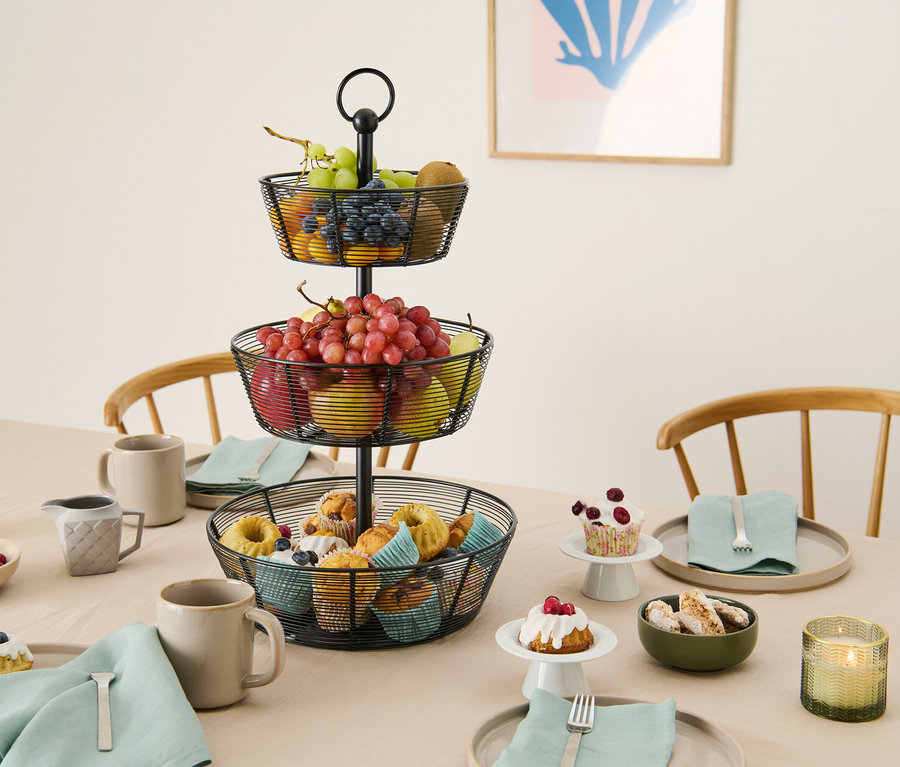 Na stole stojí třípatrový košík na ovoce z kovového drátu s pečivem, ovocem a nádobím.
