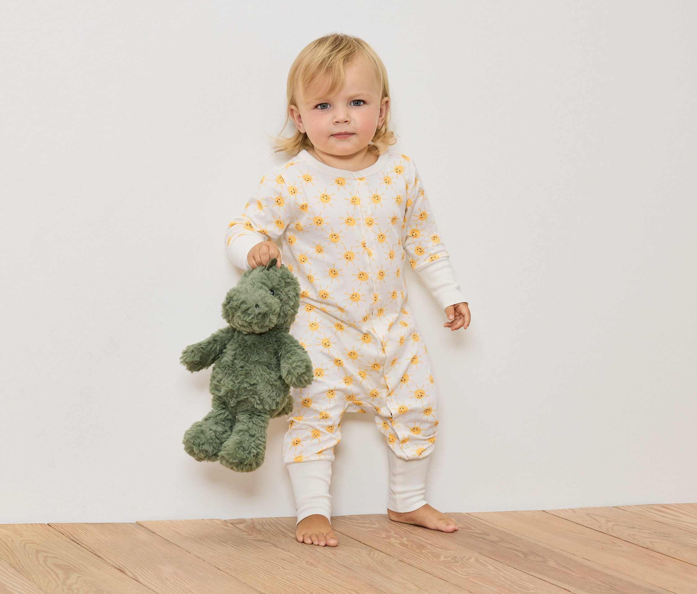 Blonďaté dítě v overalu se slunečním vzorem drží zeleného plyšového dinosaura.