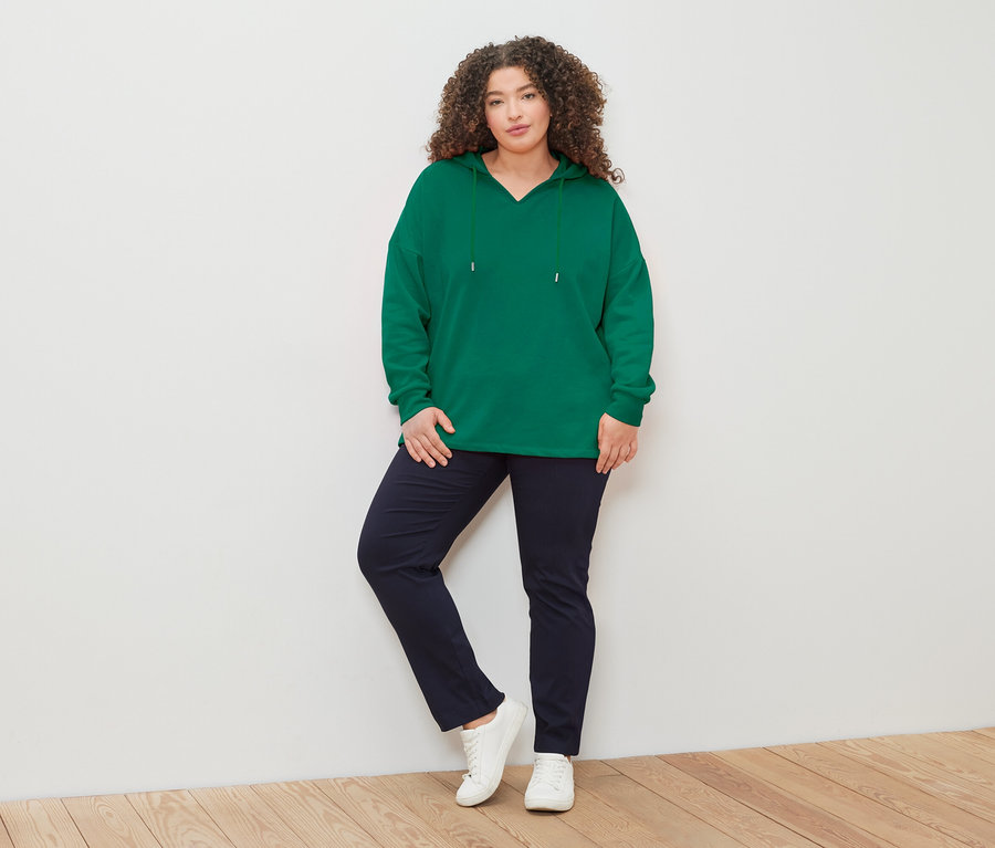 Žena s kudrnatými vlasy v zelené mikině s kapucí, tmavě modrých kalhotách a bílých teniskách pózuje před bílou zdí.
