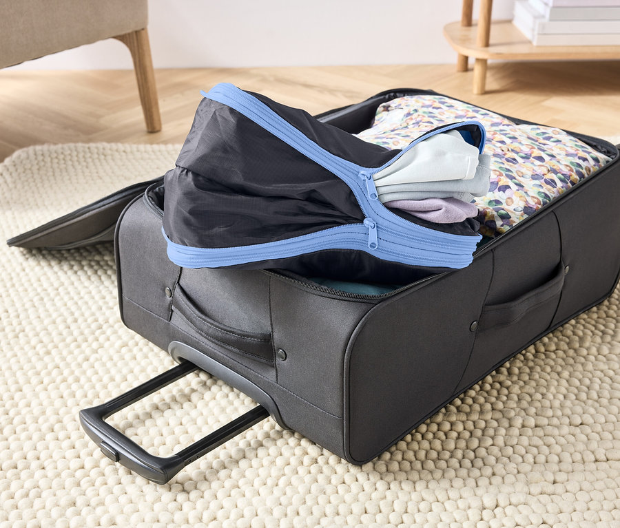 Otevřený šedý kufr na koberci, vyplněný oblečením a černou balicí kostkou s modrým zipem.