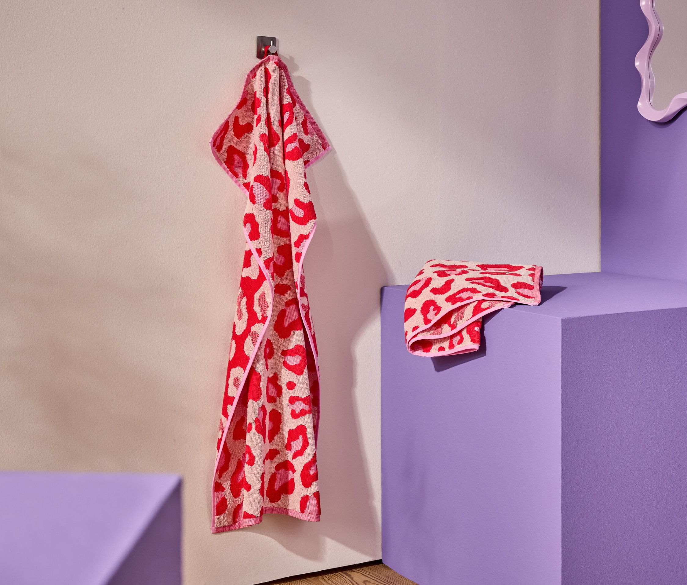 Červeno-růžový ručník s leopardím vzorem visí na háku. Složený ručník leží na fialovém podstavci.