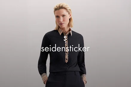 Žena s blond vlasy má na sobě černou košili s kostkovaným vzorem na límci a manžetách.