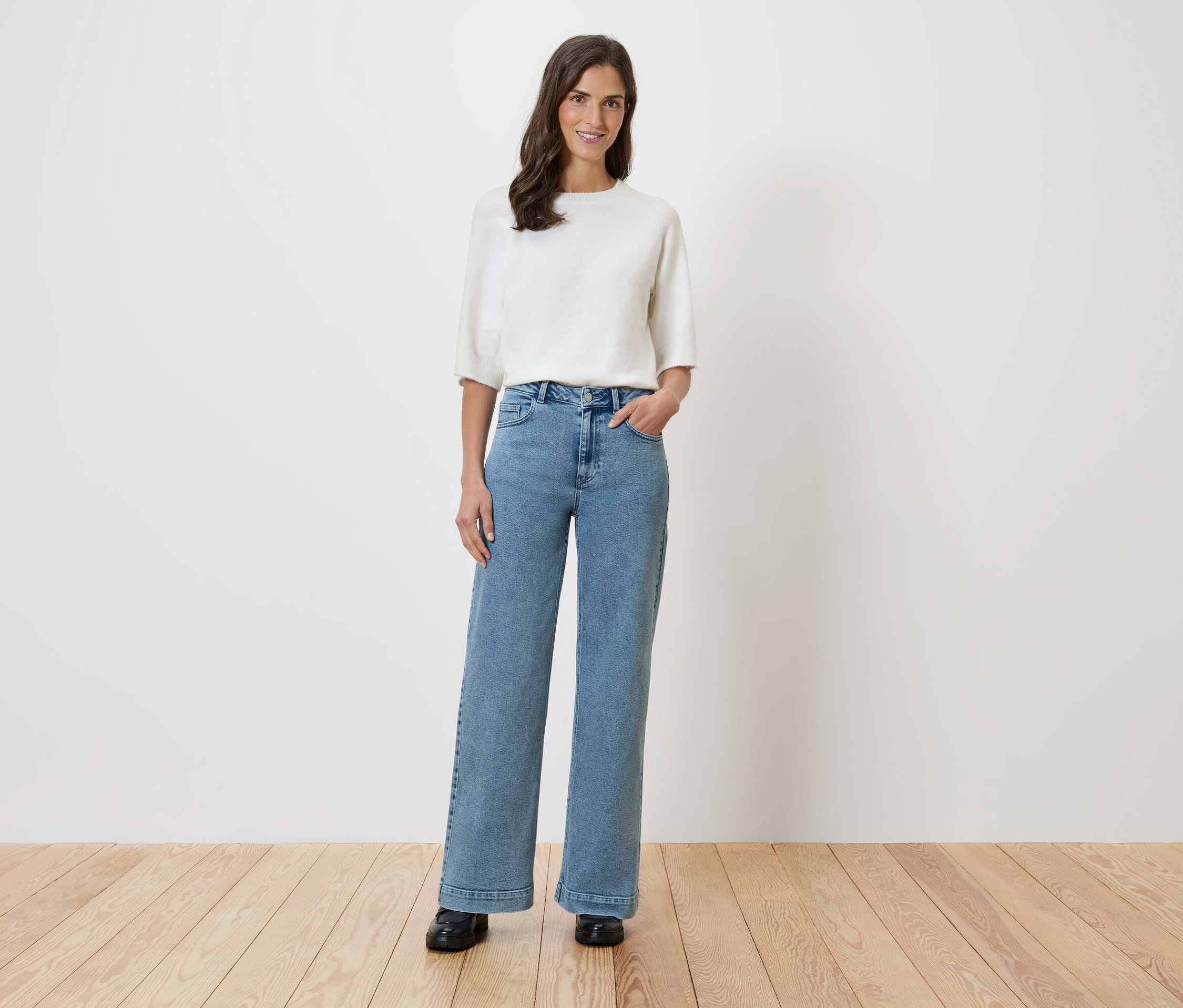 Žena stojí před bílou stěnou a má na sobě bílou halenku a široké džíny.