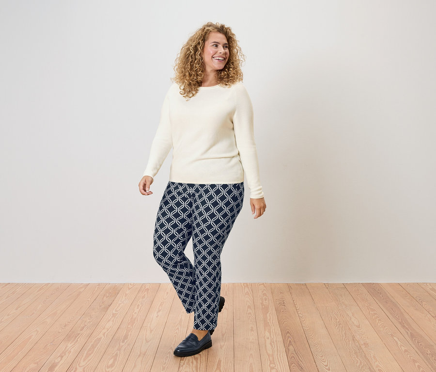 Žena s kudrnatými vlasy v krémovém svetru a tmavě modrých kalhotách s geometrickým vzorem.