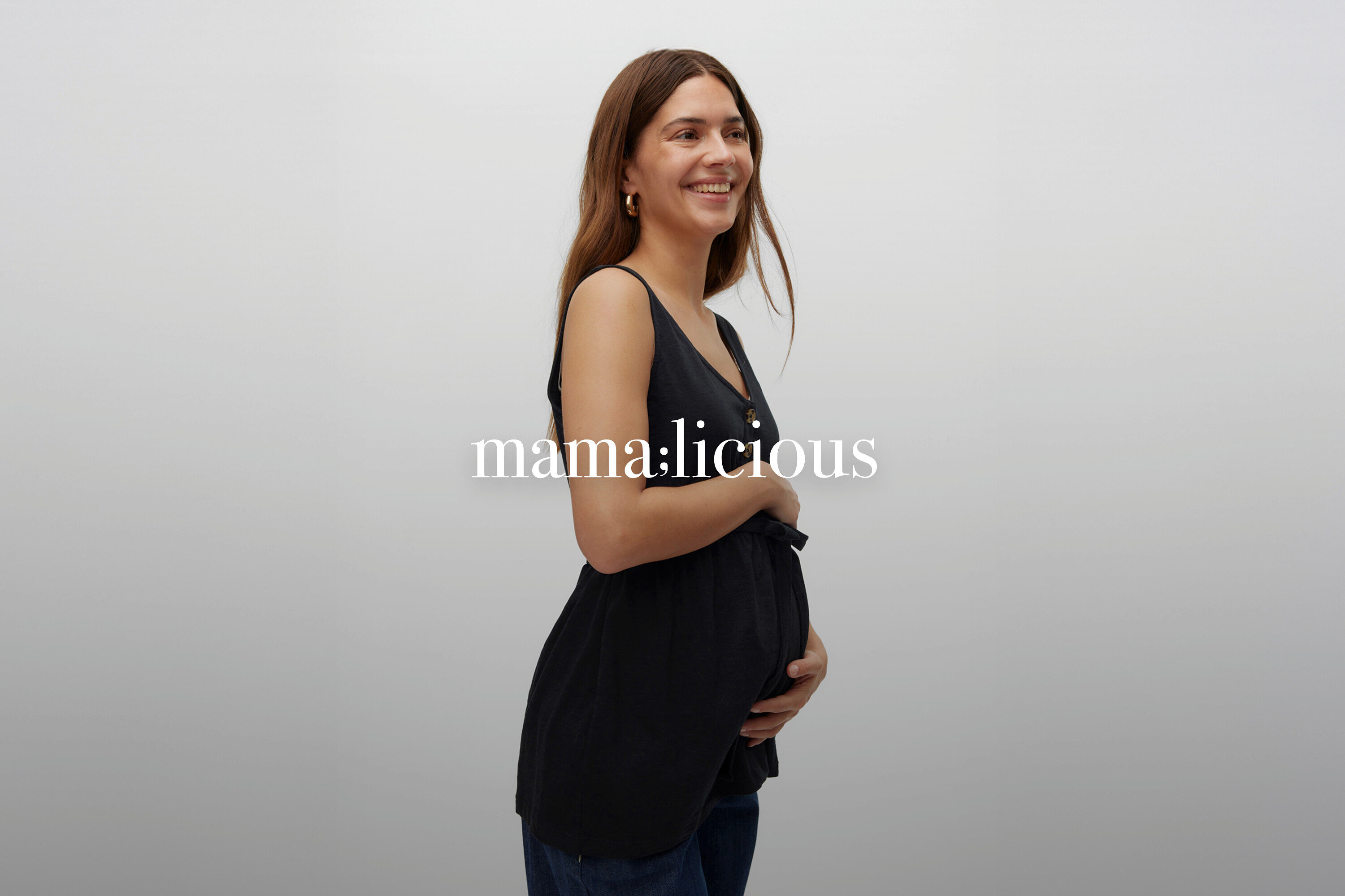 Usmívající se těhotná žena s dlouhými hnědými vlasy v černém těhotenském topu bez rukávů a džínách.