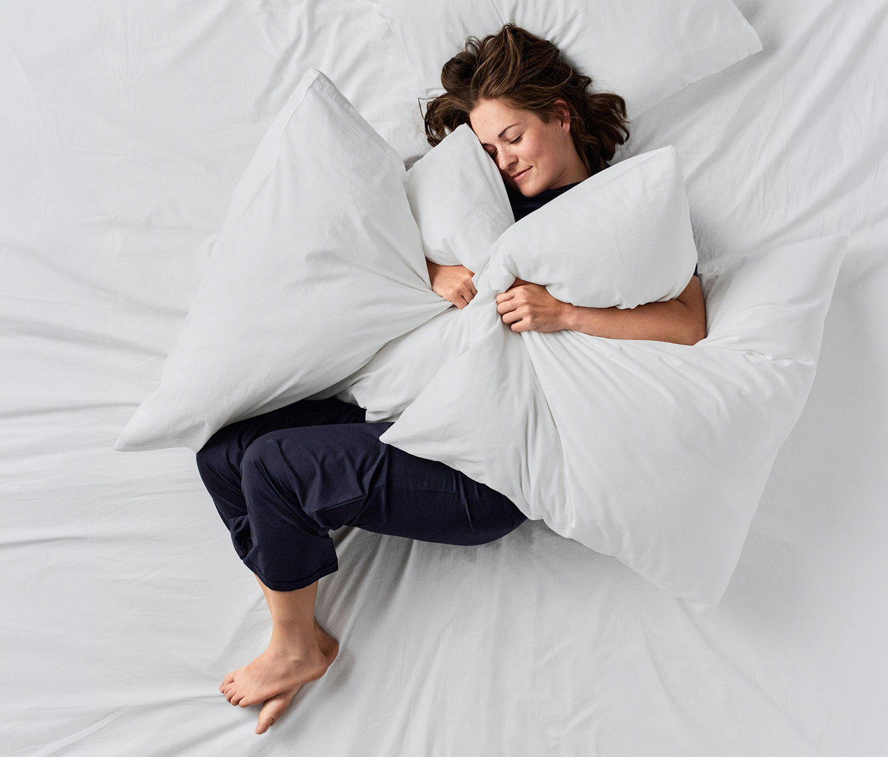 Žena spící na bílé posteli, objímající velký bílý polštář. Má na sobě tmavě modré kalhoty CALIDA »Deepsleepwear Balancing«.