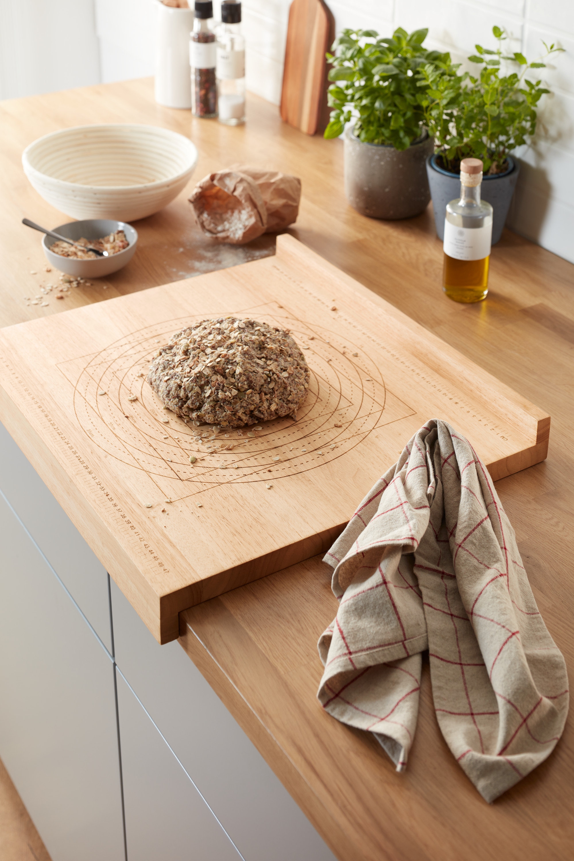 Na kuchyňské lince leží dřevěný kuchyňský vál na těsto s bochníkem chleba. V pozadí jsou bylinky v květináčích a další kuchyňské potřeby.
