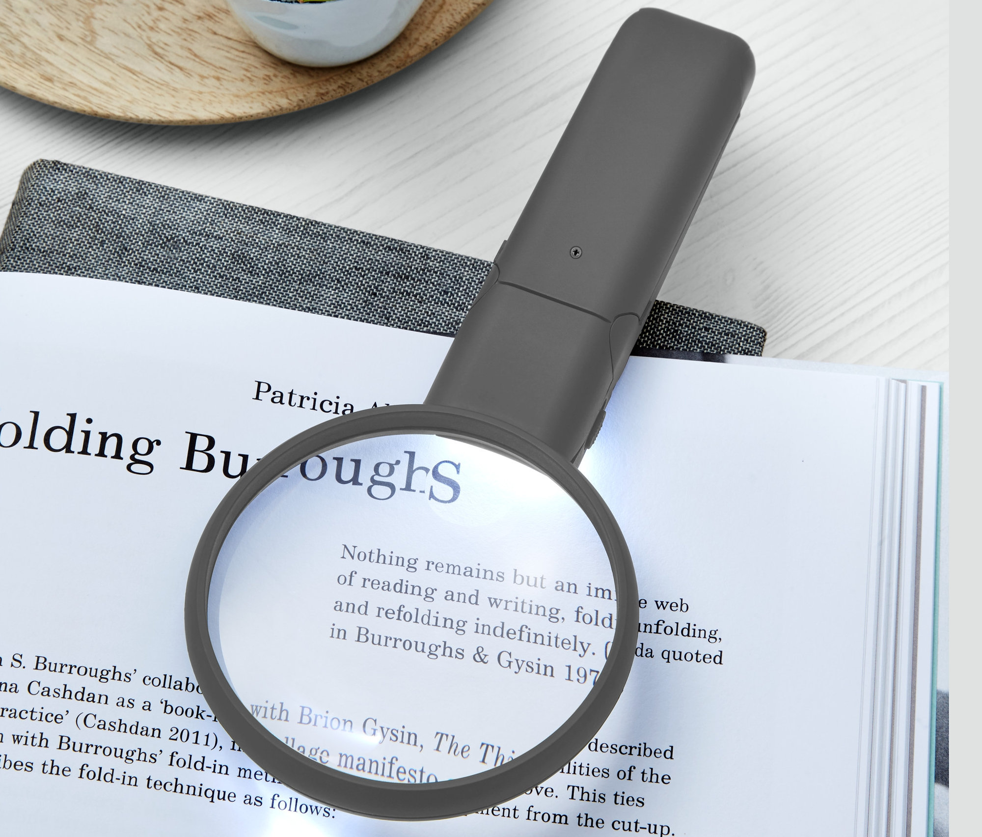 Na otevřené knize leží LED lupa na čtení a vedle ní je sukulent v bílém hrnku.