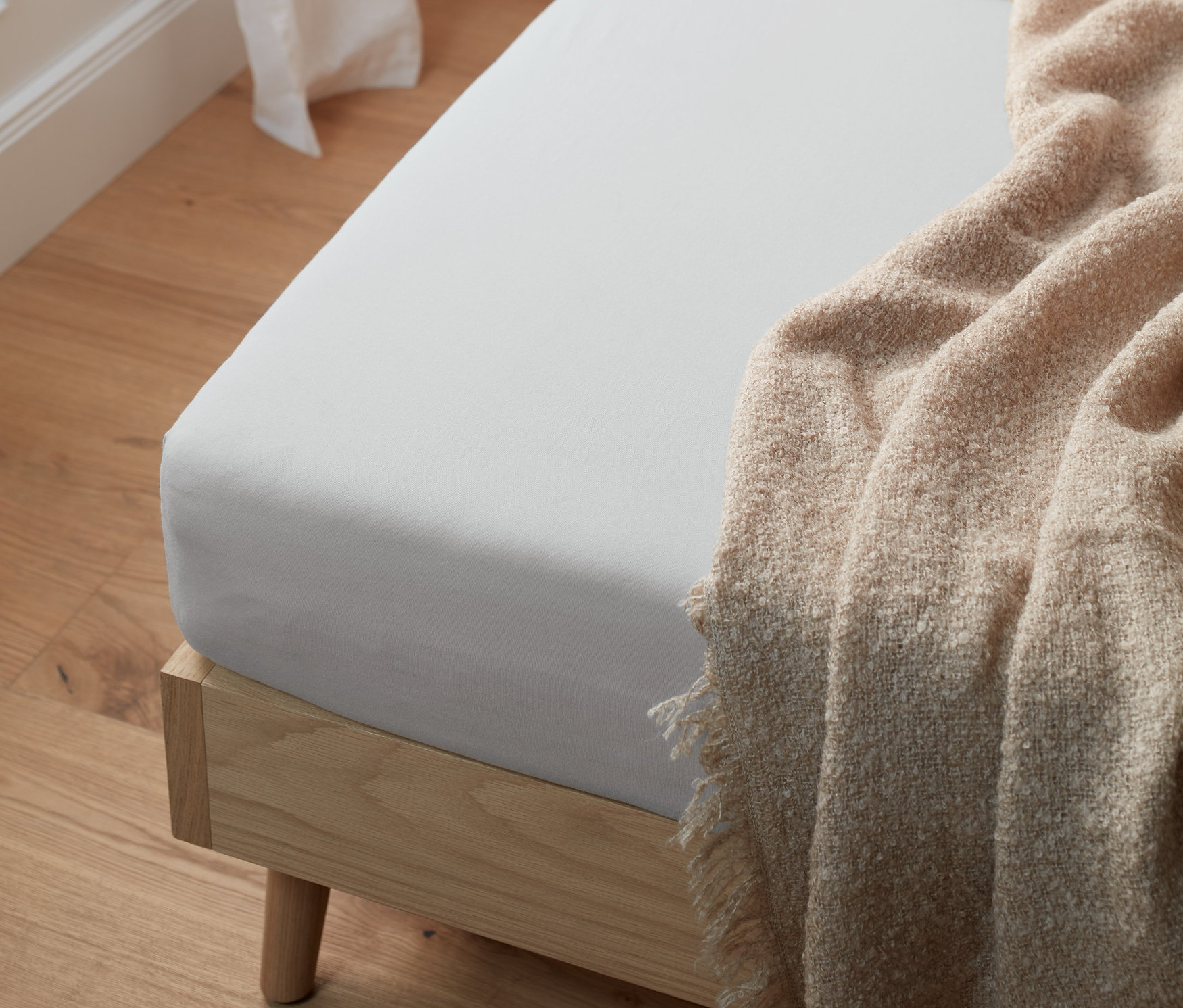 Světle šedé žerzejové napínací prostěradlo standardní velikosti na posteli s přehozenou béžovou dekou.