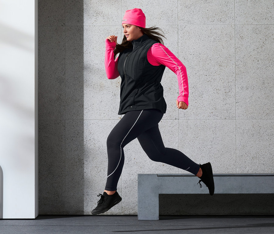 Žena v prošívané termovestě, sportovních legínách, termo čepici a neonově růžovém funkčním triku s dlouhým rukávem běží.