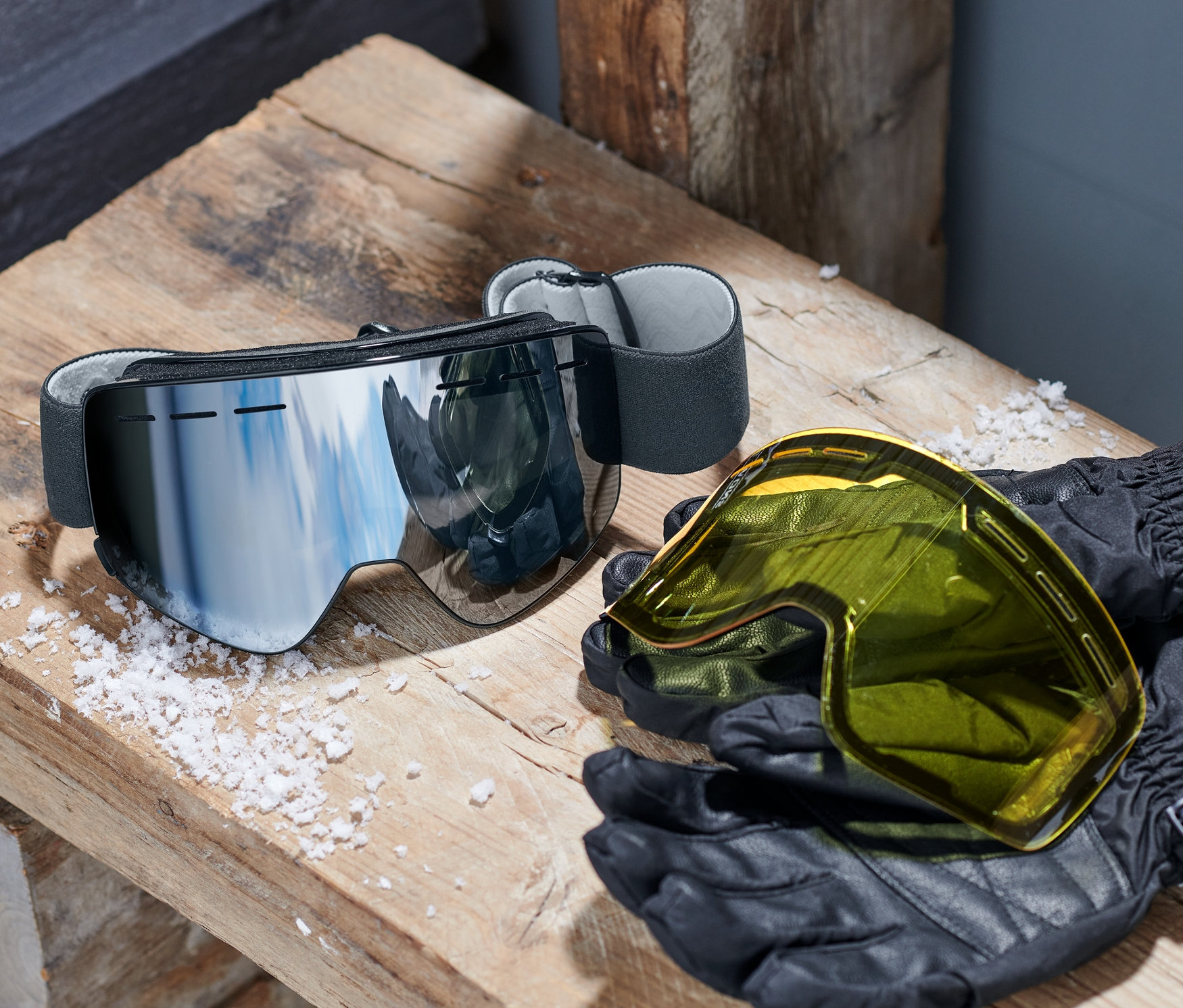 Sněhové brýle s magnetickými výměnnými zorníky leží na dřevěné lavici vedle žlutých zorníků a černých rukavic.