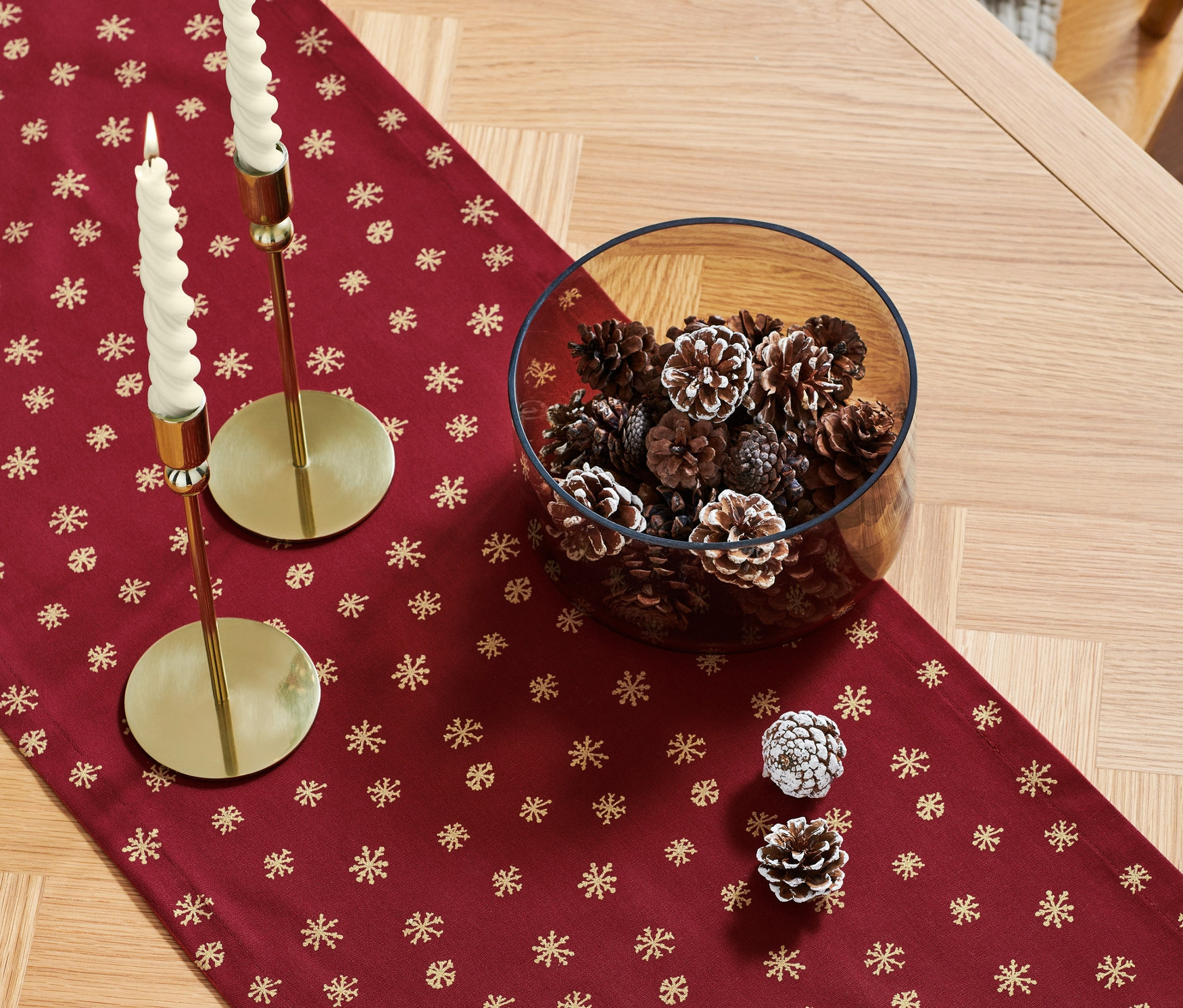 Na dřevěném stole leží červený běhoun s motivem zlatých sněhových vloček, na němž stojí dvě zlaté svícny s bílými svíčkami a skleněná mísa plná šišek.