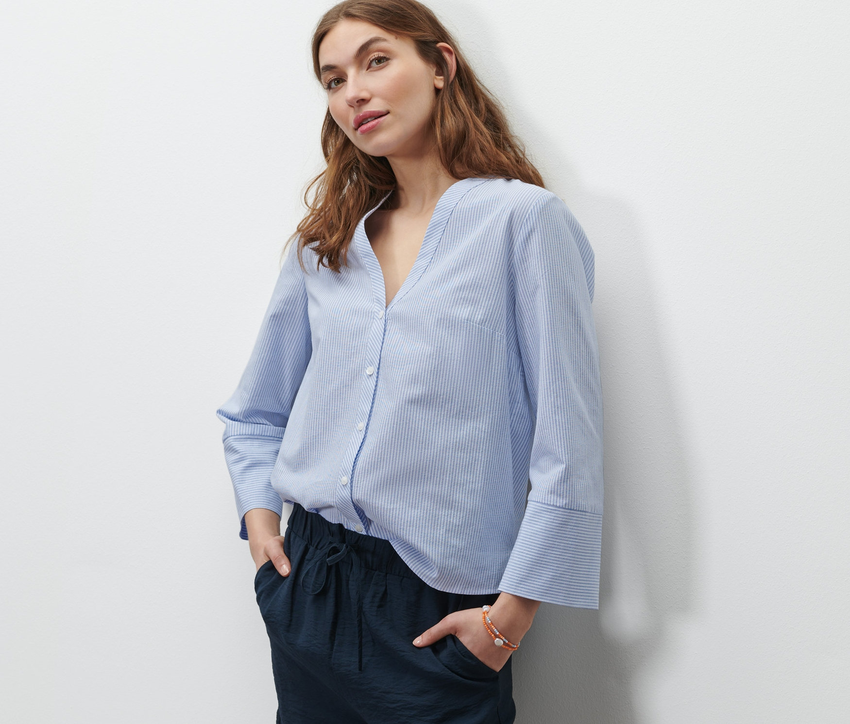 Žena stojí a má ruce v kapsách tmavě modrých šortek. Má na sobě světle modro-bílou proužkovanou halenku.