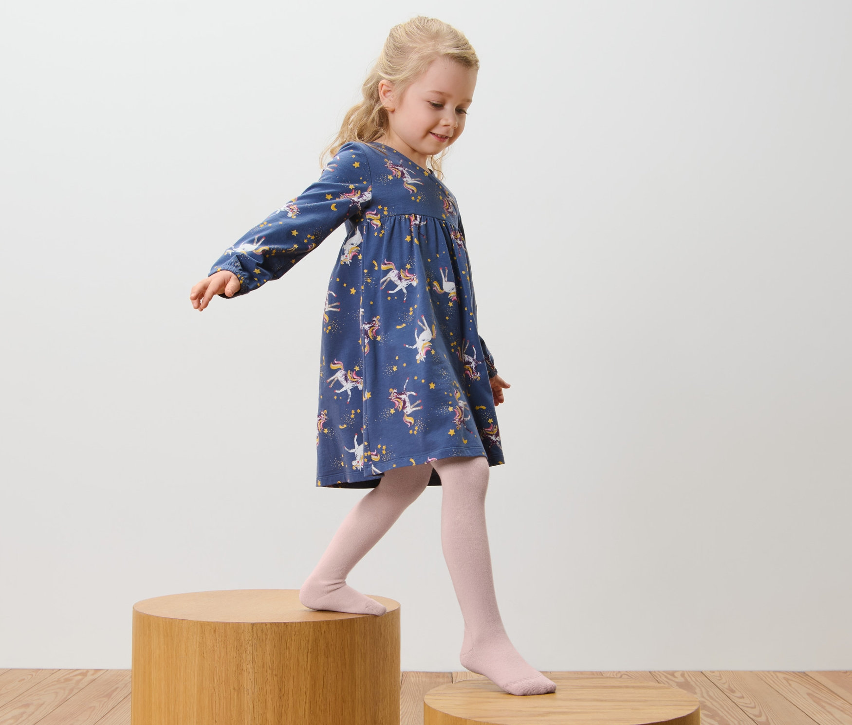 Dívka v modrých šatech s potiskem jednorožců a v růžových bavlněných punčocháčích, 5 ks, růžová/hnědá, kráčí po dřevěných stupních.