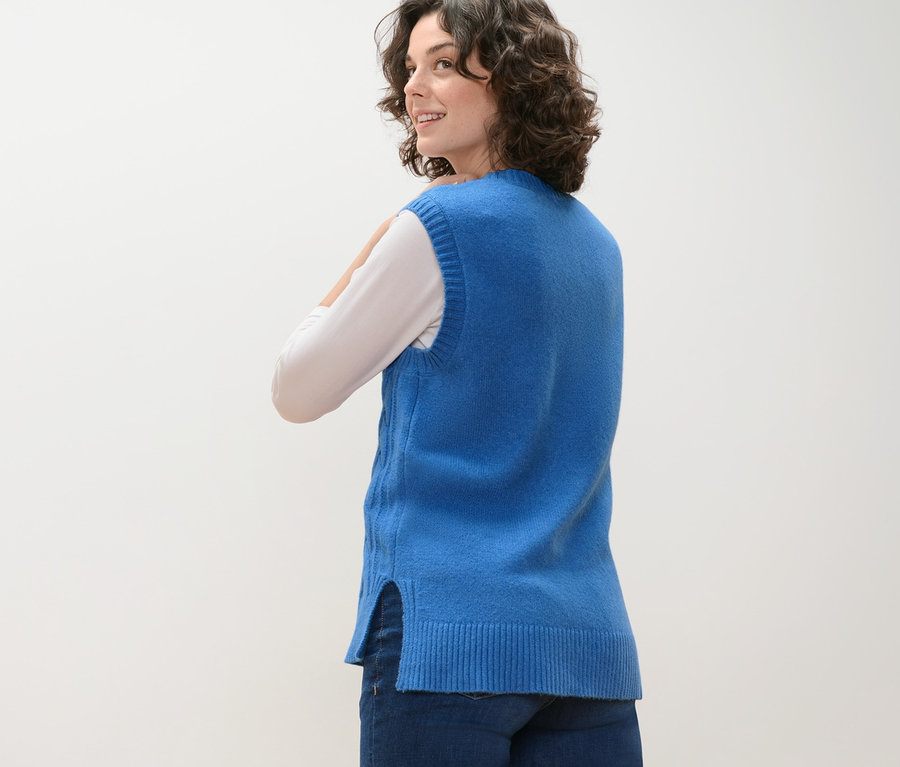 Žena se dívá přes rameno a má na sobě pletenou vestu s copánkovým vzorem.