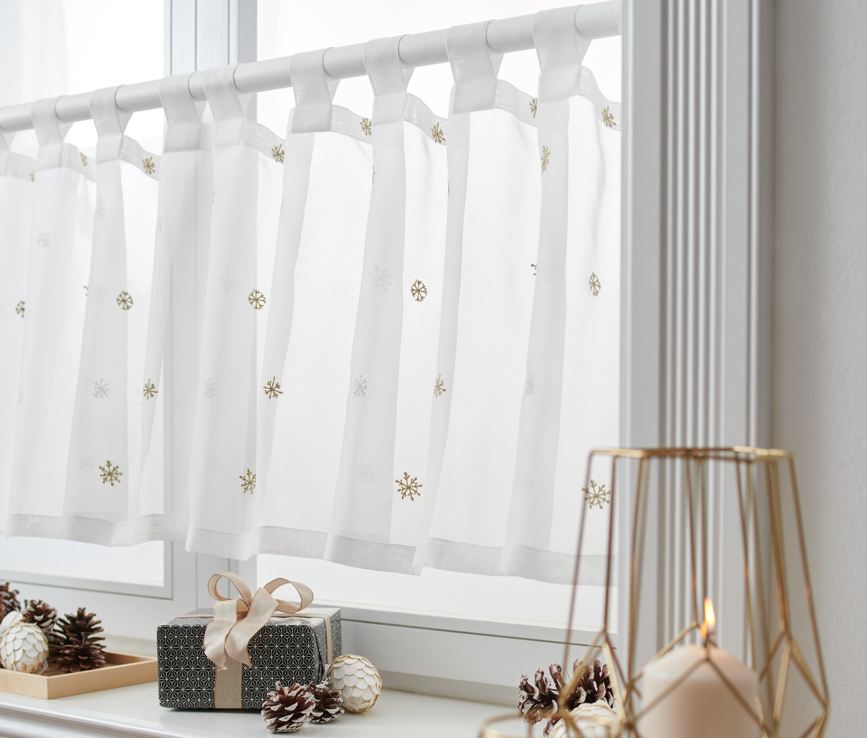 Bílá krátká záclona s motivem vloček visí v okně. Na parapetu leží dárková krabička a šišky.