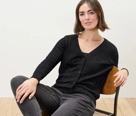 Žena sedí na židli a má na sobě černý kardigan z jemného úpletu s copánkovým vzorem.