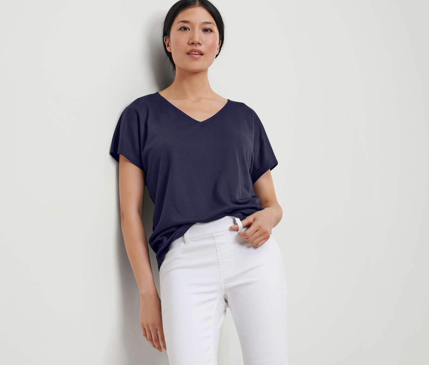 Žena opřená o zeď má na sobě tmavě modré strukturované tričko a bílé kalhoty.