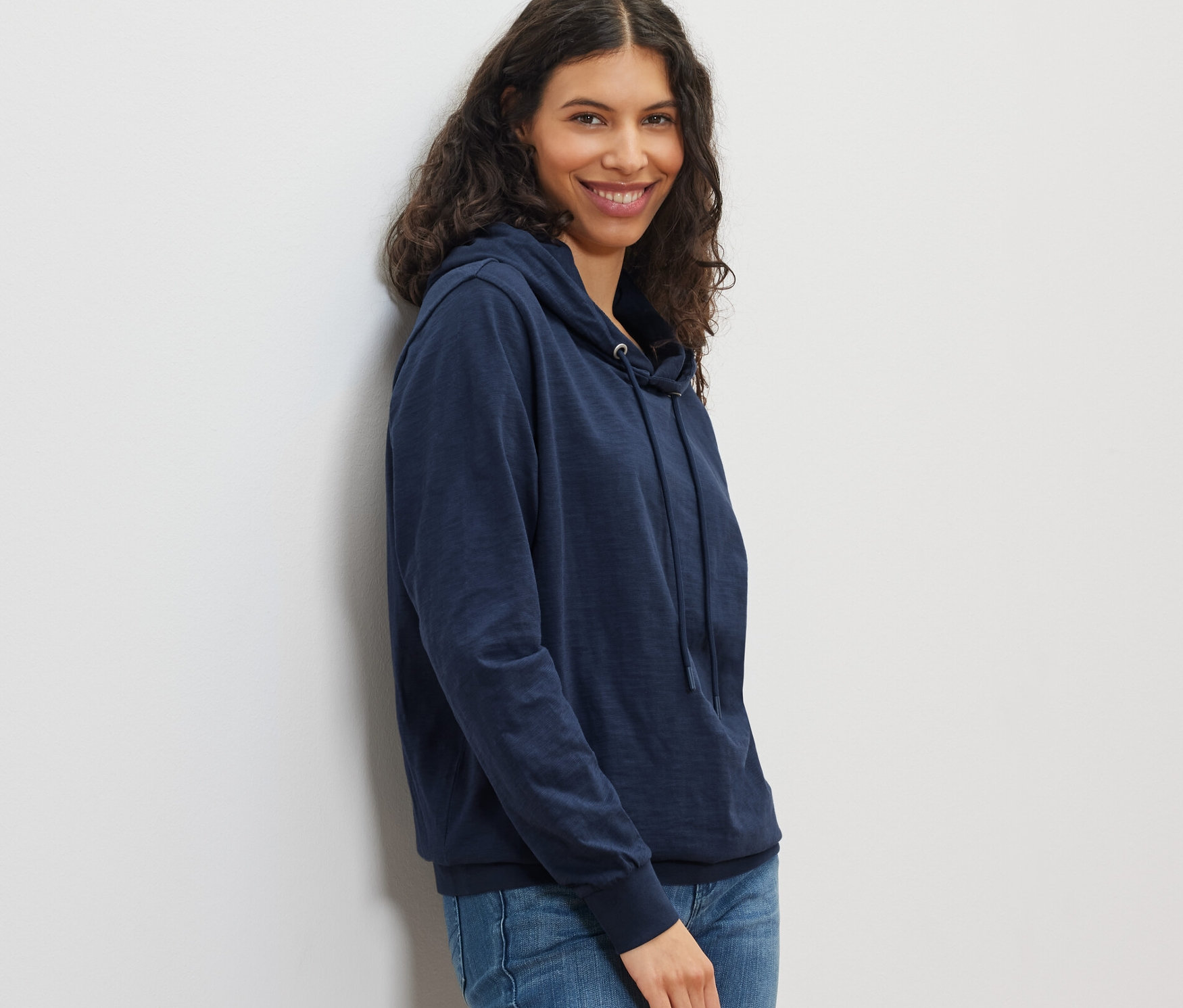 Usmívající se žena pózuje v námořnicky modré žerzejové mikině s kapucí a džínách.