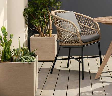 Dva jídelní křesla s polyratanovým výpletem stojí u kulatého zahradního stolu. Nad stolem visí solární filamentová závěsná lampa. Vlevo stojí květináč na kolečkách.