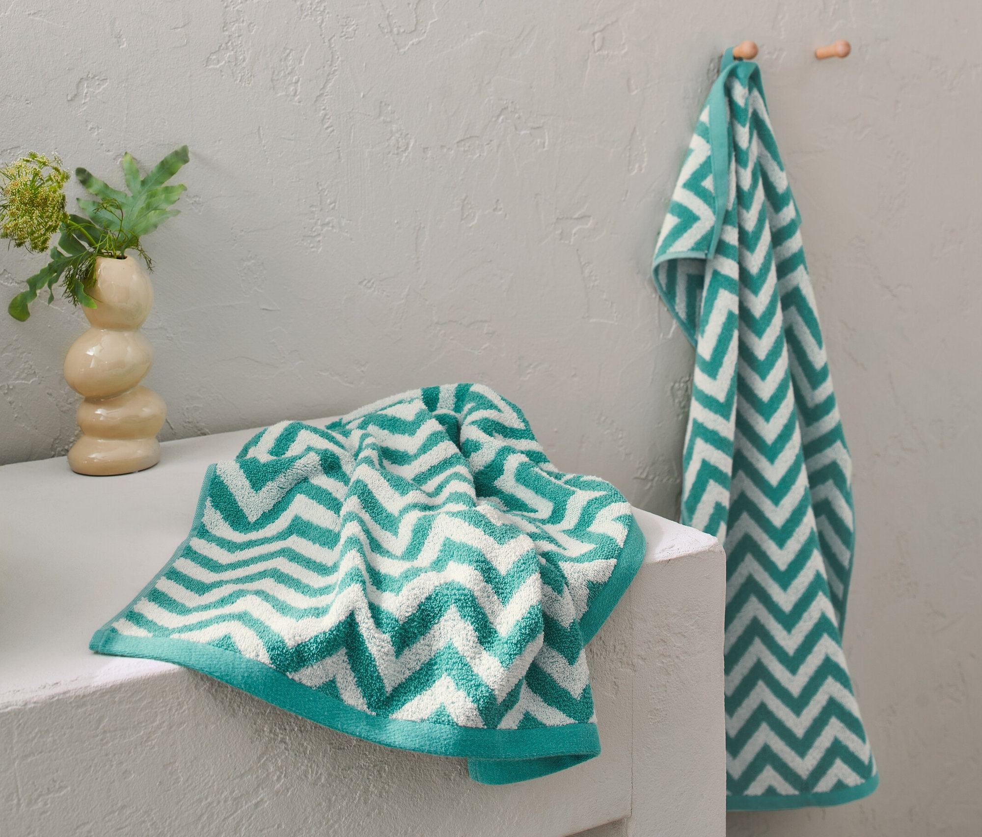 Dva žakárové ručníky modrozelené barvy leží na umyvadle, jeden visí na háčku.