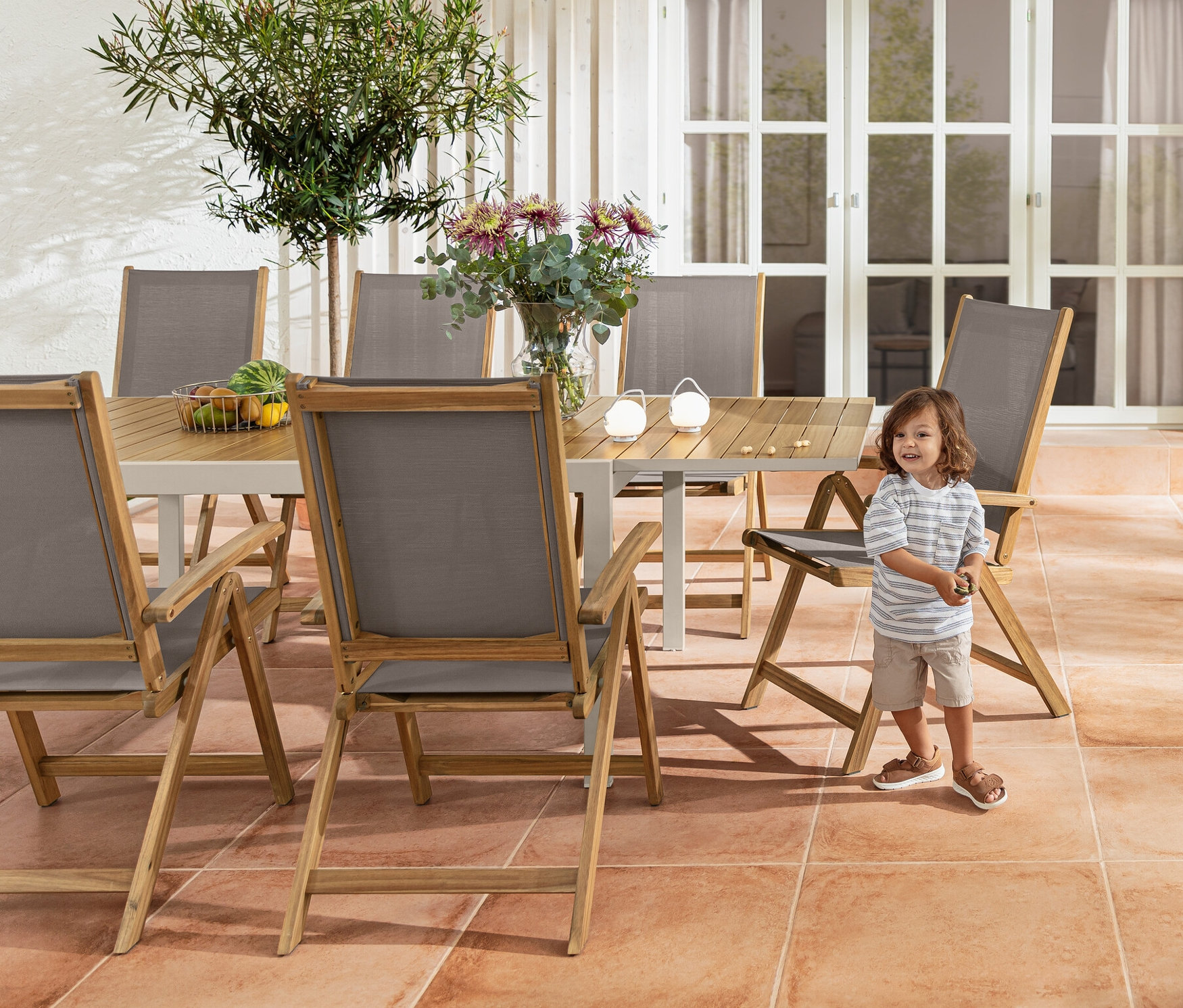 Chlapec stojí u hliníkového rozkládacího stolu »Liska« a vysokého křesla »Lenja« s dřevěnou konstrukcí. V pozadí je barový vozík a sada dobíjecích stolních lamp.