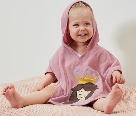 Usmívající se dítě sedí na růžovém povrchu a má na sobě froté pončo s kapucí s motivem princezny.
