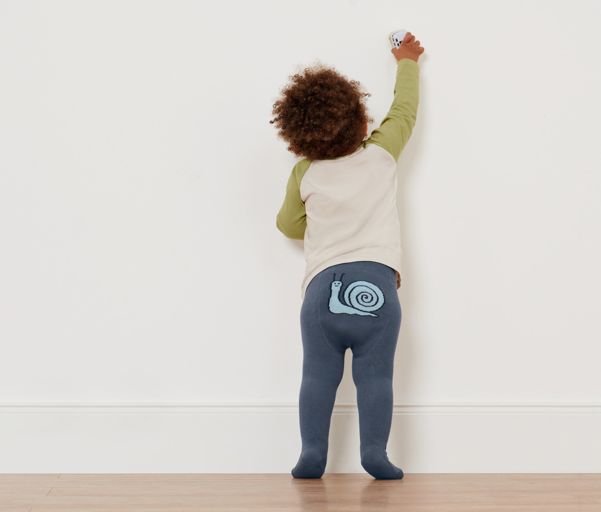 Chlapeček v punčocháčích s motivem šneka se natahuje ke zdi. Na sobě má 4 punčocháče pro miminka a malé děti.