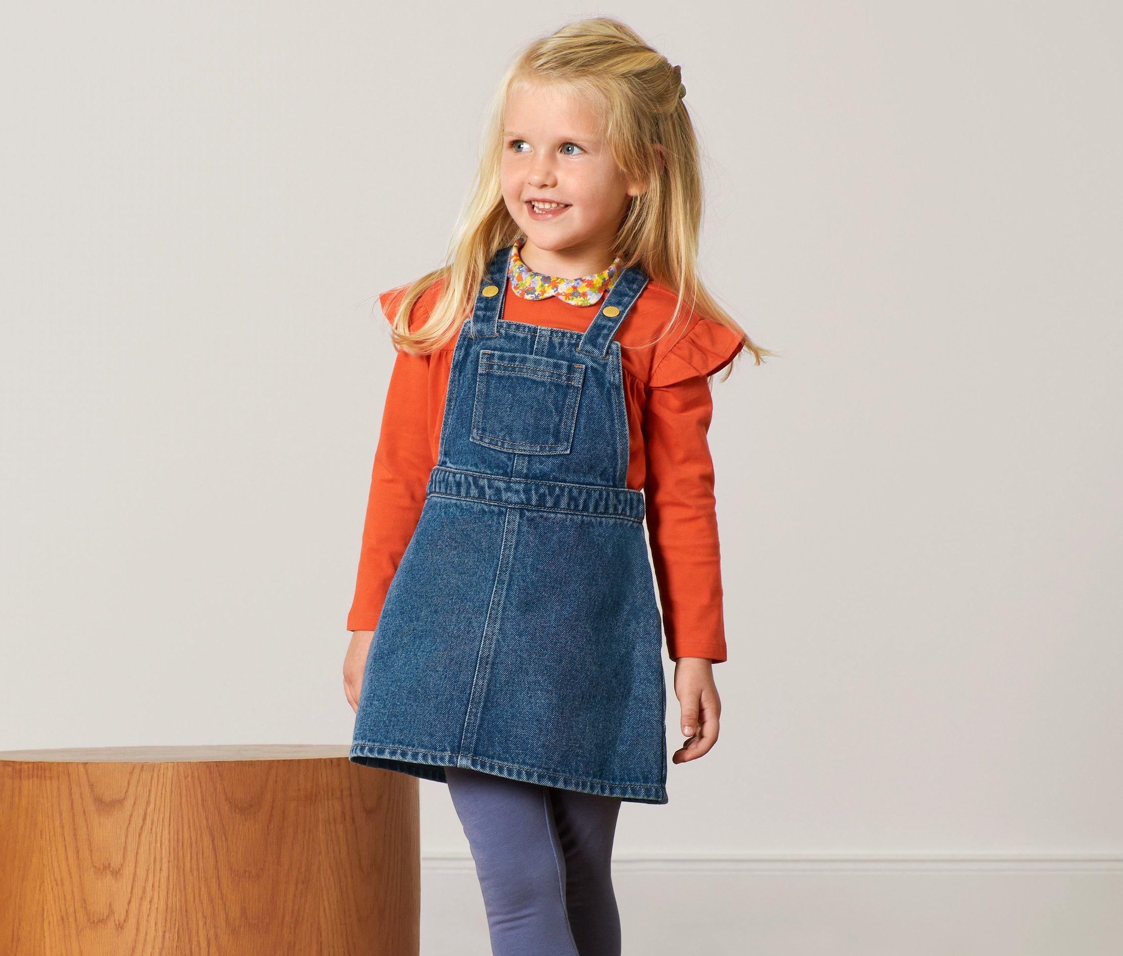 Usmívající se blondýna má na sobě džínové šaty s laclem a oranžové tričko.