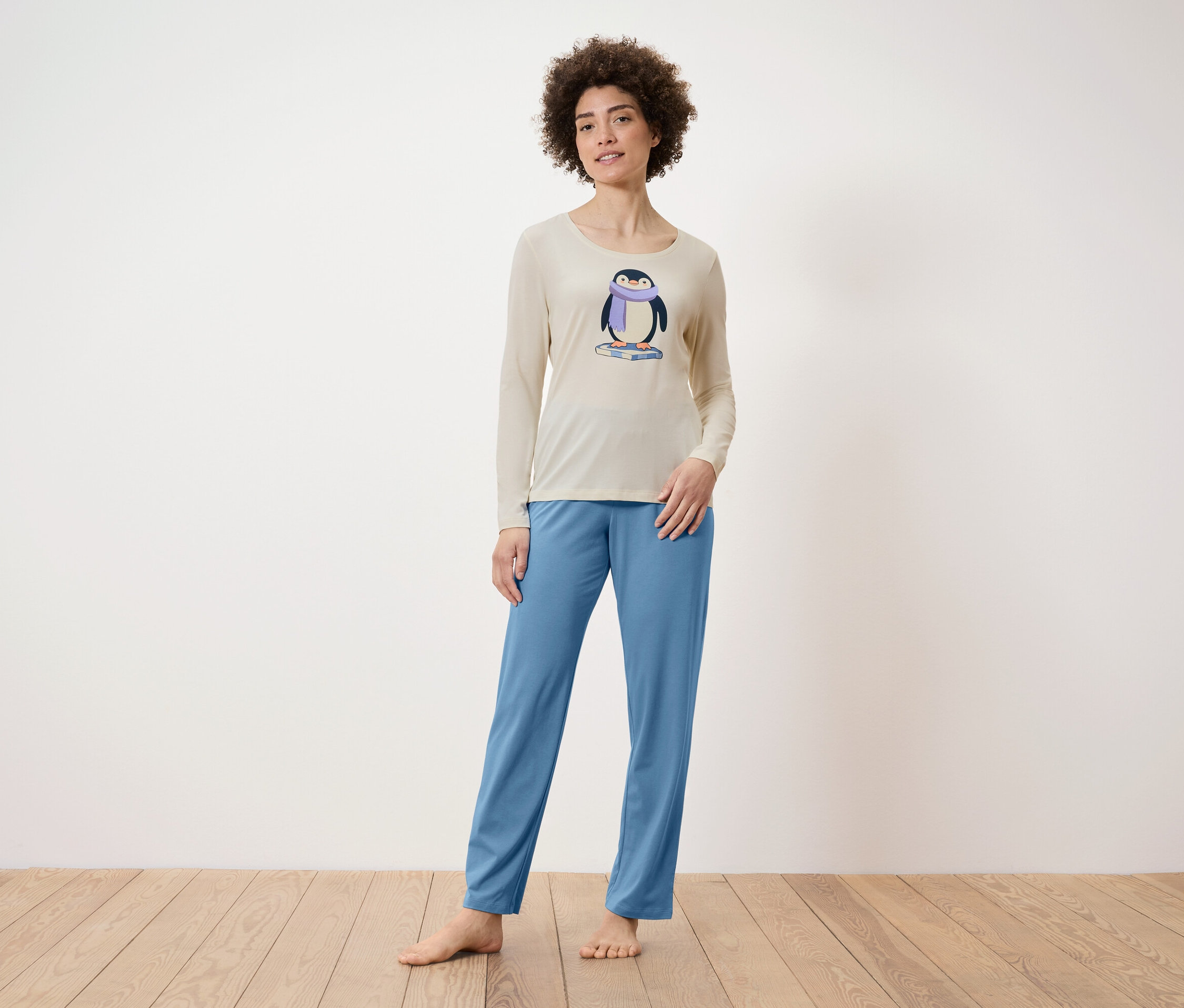Žena stojí a má na sobě béžové pyžamo s potiskem tučňáka a modré kalhoty.