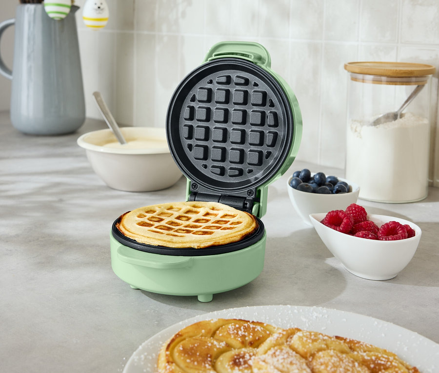 Otevřený, kulatý, mátově zelený vaflovač s vafle uvnitř stojí na kuchyňské lince. Vedle jsou dva spirálovité pečené výrobky na talíři, miska s borůvkami a miska s malinami.