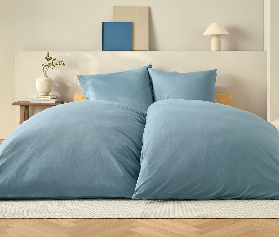 Modrá přikrývka a dva polštáře na posteli na bílém koberci.