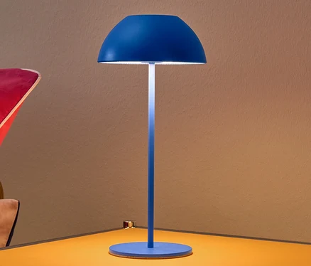 Modrá bateriová stolní lampa s LED na žlutém nočním stolku.