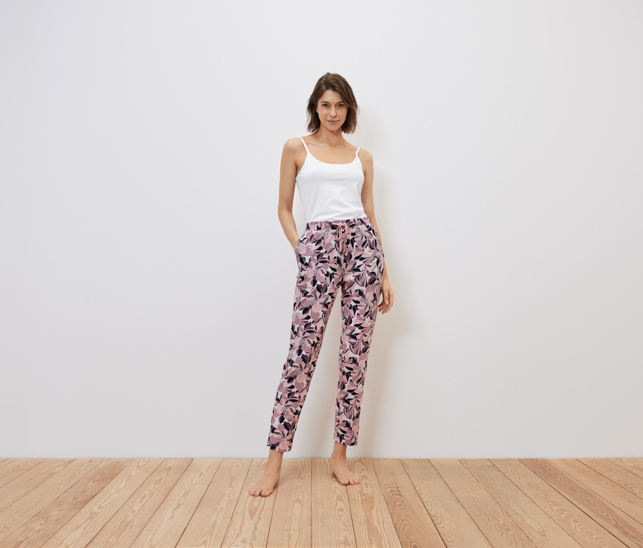 Žena stojí a má ruce v kapsách kalhot s květinovým vzorem, bílý top.
